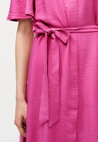 Κοντινό πλάνο ενός ροζ φόρεματος με υφή, κοντά μανίκια και ζώνη από ύφασμα που δένει στη μέση, εμφανίζοντας μέρος του χεριού και του βραχίονα ενός ατόμου.