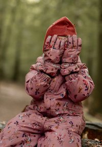 Rózsaszín virágmintás hótalpas kezeslábas, hozzáillő kesztyűvel. A gyerek az arcát takarja, piros kötött sapkát visel. A textil puha és meleg hatású.