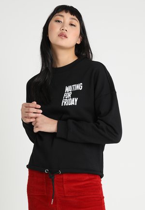 Sweatshirt noir avec "WAITING FOR FRIDAY" imprimé en blanc. Présente un col rond et un ourlet avec cordon de serrage. Porté par-dessus un pantalon en velours côtelé rouge.