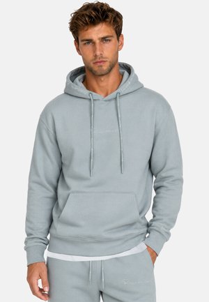 Homme aux cheveux châtain clair portant un sweat à capuche gris clair et un pantalon de jogging assorti, les mains détendues sur les côtés.