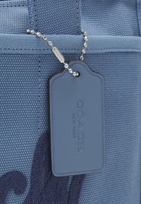 Étiquette de sac Coach bleu suspendue à une chaîne à billes argentée attachée à un tissu bleu texturé avec des bords cousus.
