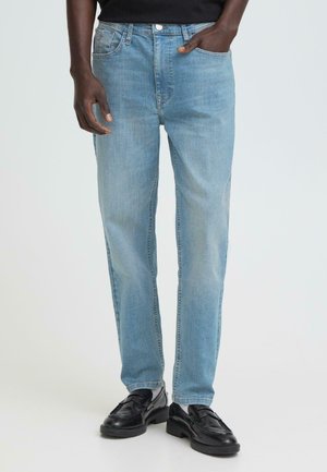 Mand iført lyseblå jeans med lige ben, sorte læderloafers og en sort skjorte, hånd i lommen, står på hvid baggrund.