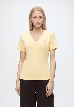 Femme aux cheveux blonds raides porte un T-shirt jaune clair à col en V et un pantalon foncé, debout devant un fond blanc uni.