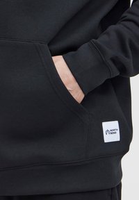 Čierna mikina s kapucňou vyrobená z mäkkej tkaniny, ktorá má klokaní vrecko a rebrované manžety. Na prednej strane je biely štítok s logom "NORTH BEND".