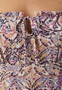 Vzorovaný textilní top s volánkovým výstřihem, který má detaily ve tvaru klíčové dírky a tkaní. Barvy zahrnují béžovou, levandulovou, růžovou a černé akcenty.