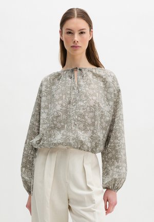 Woman wearing a loose, long-sleeve floral blouse with a keyhole neckline and high-waisted cream pleated trousers.