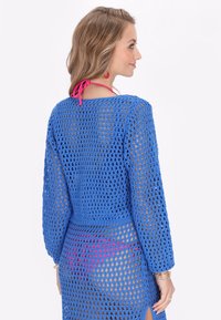 Kék, horgolt kötésű, hosszú ujjú takaró, nyitott szövésű dizájnnal. Karcsúsított derékkal és oldalsó hasítékkal rendelkezik, amit egy rózsaszín kötés díszít.