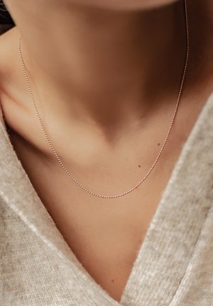 Dünne Silberkettnecklace mit kleinen, runden Perlen, die auf dem Schlüsselbein ruhen und einen hellen, strukturierten Pullover ergänzen.