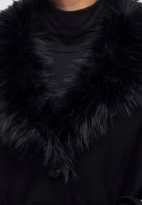 Gros plan d'un manteau noir avec un grand col en fourrure noire et duveteuse porté sur un haut noir.