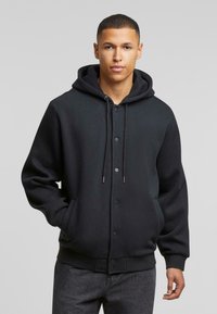 Zwarte hoodie met voor knopen, trekkoorden en zijzakken. Zachte textuur, relaxte pasvorm en ribgebreide manchetten en tailleband.