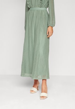 Femme portant une longue robe en dentelle verte sauge à manches longues et ceinture, associée à des sandales blanches à talons et bouts ouverts.