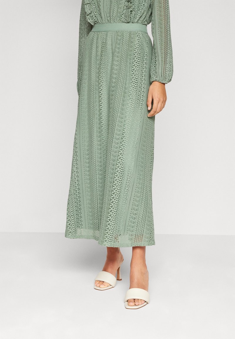 Femme portant une longue robe en dentelle verte sauge à manches longues et ceinture, associée à des sandales blanches à talons et bouts ouverts.