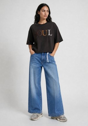 Woman standing with hands in pockets wearing black t-shirt with "SOUL" text, wide-leg blue jeans, and gray sneakers on gray background.