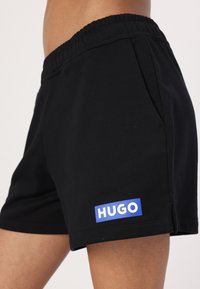 Juodi medvilniniai šortai su elastingu juosmeniu, šoninėmis kišenėmis ir mėlynu "HUGO" logotipu kairėje apatinėje pusėje.