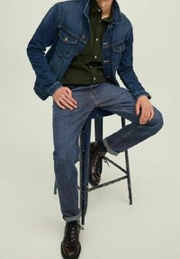 Chaqueta de mezclilla con bolsillos, camisa verde, jeans azules y botas marrón oscuro, sentado en un taburete contra un fondo neutro.