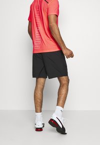 Kirkas koralli urheilupaita, jossa on "PUMA" -grafiikka, yhdistetty mustiin shortseihin. Valkoiset tennarit punaisilla yksityiskohdilla ja valkoiset sukat. Minimalistinen tausta.