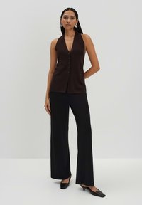 Top senza maniche marrone con scollatura a V profonda e dettagli con bottoni, abbinato a pantaloni neri a gamba larga e scarpe con tacco nere.