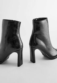Bottines en cuir noir brillant avec talons blocs et fermetures éclair latérales, vues de dos et de profil sur un fond uni clair.