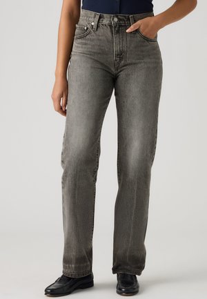 Personne portant un jean gris délavé à jambes droites avec une taille haute, une main dans la poche, associé à des chaussures noires pointues sur un fond uni.