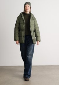 Giacca imbottita verde con cappuccio, indossata sopra un maglione scuro, abbinata a jeans blu larghi e scarpe marroni; berretto verde a coste sulla testa.