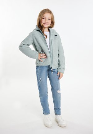 Felpa grigia chiara con zip e tessuto strutturato, con logo frontale e cordini. Abbinata a jeans blu strappati e sneaker bianche.