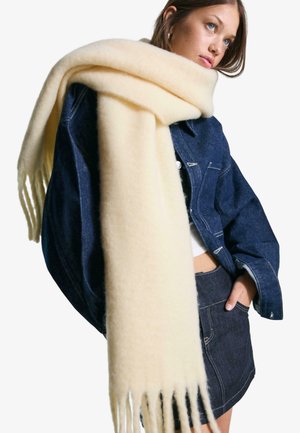 Écharpe en cream doux tricotée avec des extrémités frangées, drapée sur une veste en denim foncé. L'écharpe a une texture lisse et une apparence épaisse et confortable.