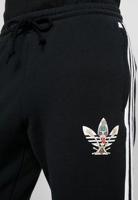 Svarta träningsbyxor med vita sidostråkar, med en detaljerad flerfärgad grafisk logotyp på övre låret. Materialet verkar mjukt och texturerat.