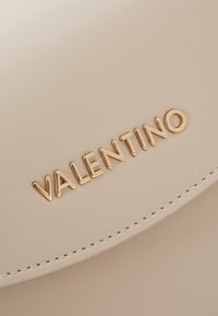 Krämfärgad läderhandväska med guld "VALENTINO"-logotyp. Har en slät textur och kontrasterande vita sömmar längs kanten.