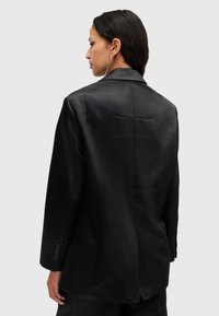 Blazer nero su misura con taglio dritto e finitura satinata. Presenta un colletto a scollo classico e polsini abbottonati. Dettagli di cucitura discreti.