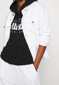 Giacca di jeans bianca con hardware argentato indossata sopra un hoodie nero con tasca frontale e testo del logo bianco. Il tessuto appare liscio e casual.