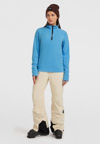 Lyseblå fleecepullover med halv lynlås, parret med beige snebukser med sorte detaljer og vandtætte støvler. Brun strikket hue inkluderet.
