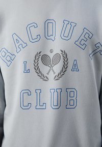 Sudadera gris claro con texto en azul y gris. Presenta un diseño de "RACQUET CLUB", raquetas de tenis y motivos de laurel. Tejido suave y texturizado.
