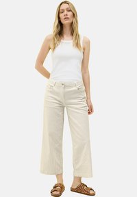 Helle beige, weit geschnittene Kordhosen mit vertikalen Streifen, kombiniert mit einem weißen Tanktop und braunen Slidesandalen mit Punkten.