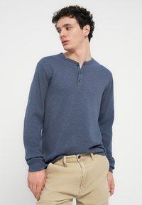 Μπλε μακρυμάνικο πουκάμισο Henley με τρία κουμπιά, υφασμάτινη υφή και στρογγυλή λαιμόκοψη, σε συνδυασμό με ανοιχτόχρωμο παντελόνι.