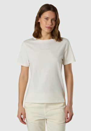 Donna con capelli castani ondulati che indossa una maglietta bianca a maniche corte e pantaloni crema, in piedi davanti a uno sfondo grigio chiaro.