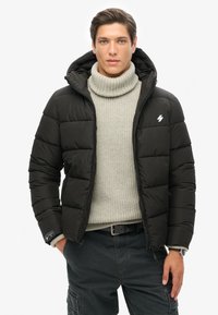 Sort pufferjakke med lynlås foran og hætte. Har en grå strikket sweater under, cargo bukser og en logo detalje på brystet.