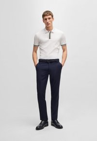 Biała koszulka polo z kołnierzykiem w czarne, granatowe i czerwone paski, zestawiona z granatowymi spodniami i czarnymi butami wizytowymi. Model stoi z rękami w kieszeniach.