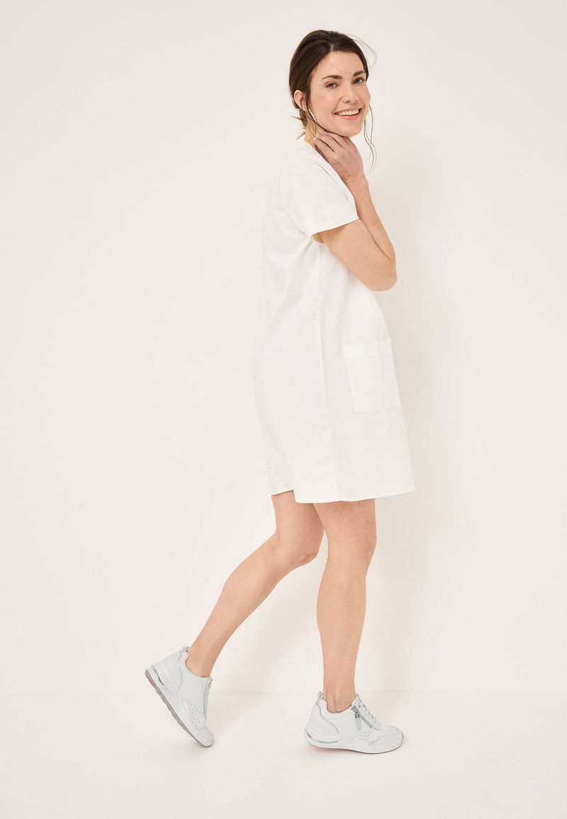 Abito di cotone bianco con maniche corte, due tasche laterali e una vestibilità rilassata. Il modello indossa sneakers grigie chiare con dettagli testurizzati.