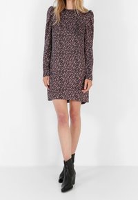 Femme portant une robe mini à manches longues rose et noire à motifs, associée à des bottines noires, se tenant devant un fond blanc uni.