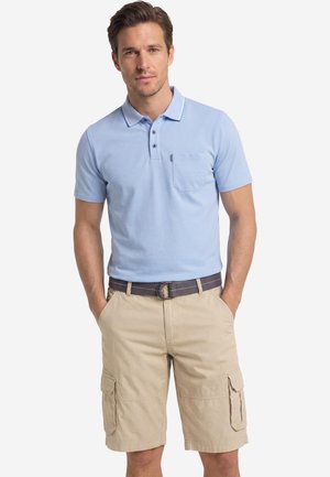 Man wearing light blue short-sleeve polo shirt and beige cargo shorts with dark belt, standing with hands in pockets against white background.