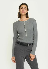 Grauer gerippter Pullover mit einem gewellten Halsausschnitt und Knöpfen vorne. Tragen mit einem schwarzen Gürtel und grauen strukturierten Hosen. Lange Ärmel.