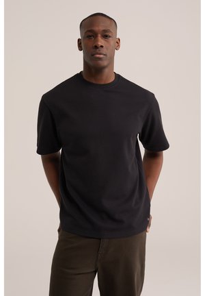 Mand står med hænderne bag ryggen iført en ensfarvet sort kortærmet t-shirt og brune bukser mod en beige baggrund.