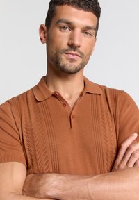 Bruine gebreide polo met een gestructureerd patroon en korte mouwen. Beschikt over een klassieke kraag en drie knopen aan de voorkant.