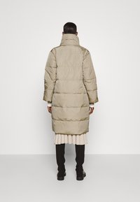 Polstret lang frakke i lys beige med højt krave, lige snit og elastiske manchetter. Glat tekstur med syede paneldetaljer.