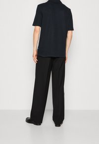 Marineblaues Polohemd mit kurzen Ärmeln und einer strukturierten Oberfläche; kombiniert mit locker sitzenden schwarzen Hosen und klobigen schwarzen Schuhen.