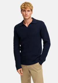 Maglione lavorato a maglia blu navy con un motivo testurizzato, scollo a V e colletto. Maniche lunghe, design aderente, abbinato a pantaloni beige chiaro.