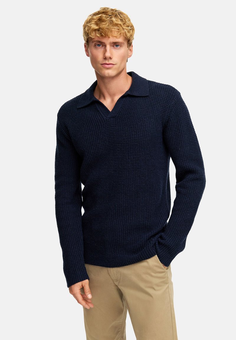 Maglione lavorato a maglia blu navy con un motivo testurizzato, scollo a V e colletto. Maniche lunghe, design aderente, abbinato a pantaloni beige chiaro.