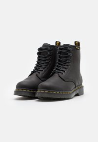 Svarte skinnankleboots med solide snørebånd, rund tå, teksturert overflate, gule sømmer langs sålen og hjelpesløyfe bak.