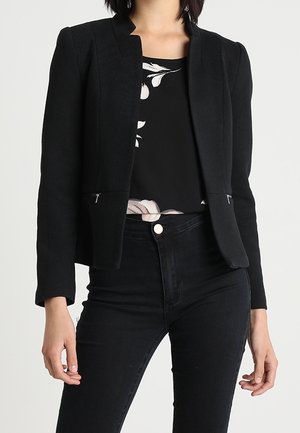 Femme portant un blazer noir ajusté sur un haut noir fleuri et un jean noir taille haute sur un fond uni.