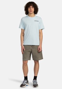 Light blue cotton t-shirt with short sleeves, featuring a small black logo. Paired with brown shorts and black socks, standing in neutral pose.